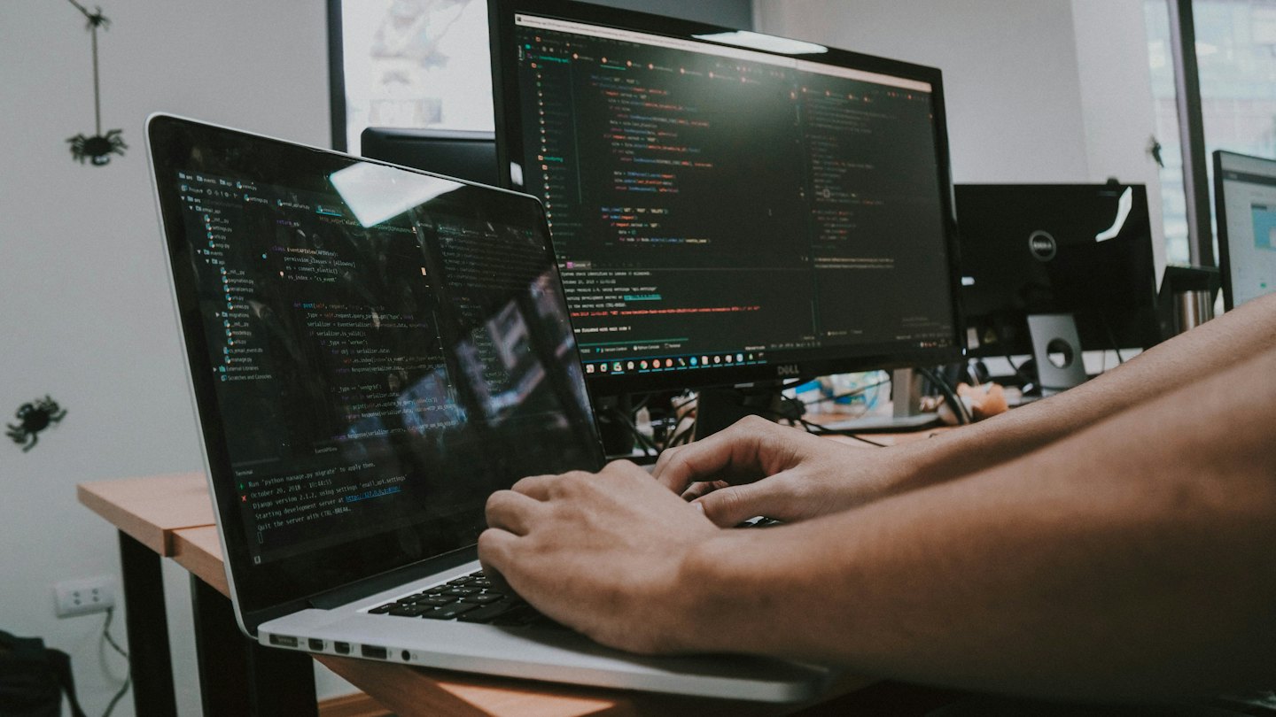 Mani che scrivono codice su un laptop collegato a un grande monitor.